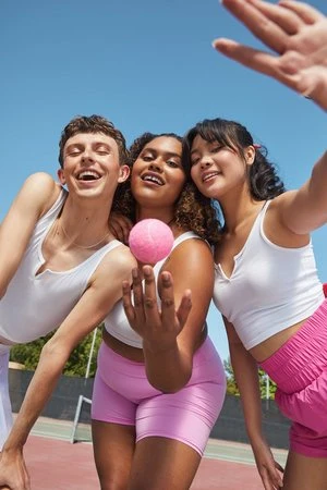 Three cheerful young adults in white tops and pink shorts holding a pink tennis ball against a clear blue sky