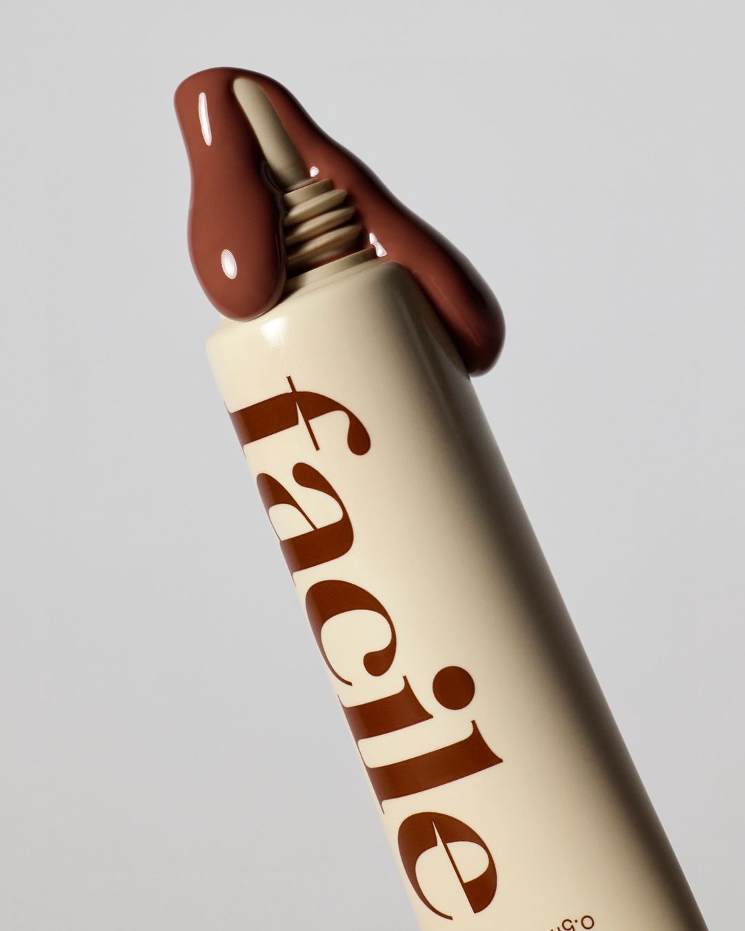 Cosmetic product in a white and beige squeeze tube, featuring brown text and a drop of brown cream, set against a light gray background.