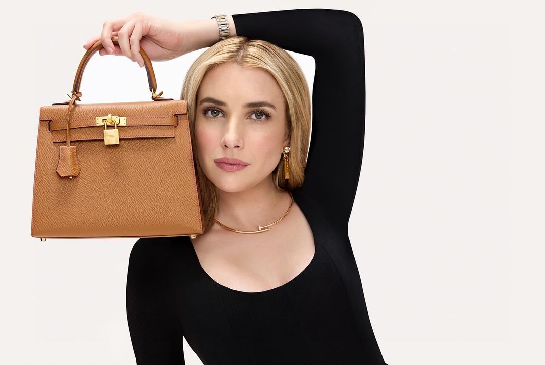 Blond woman holding a tan leather handbag with gold hardware against a white backdrop, wearing a black dress and gold jewelry.
