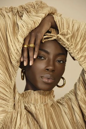 Portrait of a Black woman in a pleated gold top with hands framing her face, wearing gold hoop earrings and rings, by photographer Claire Leahy