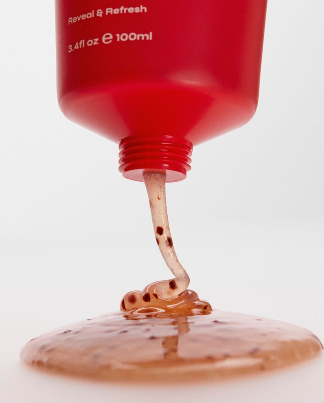 Close-up of viscous scrub flowing from a red squeeze tube onto a white surface. The scrub contains small brown particles.