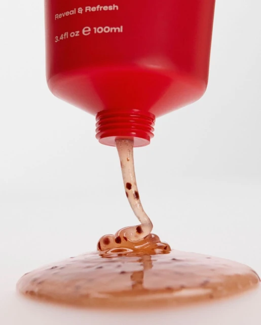 Close-up of viscous scrub flowing from a red squeeze tube onto a white surface. The scrub contains small brown particles.