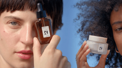 Two women showcase skincare products against a light blue backdrop, featuring a serum bottle and a moisturizer jar in their hands.