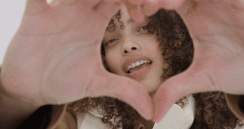 Smiling woman with curly hair forms heart shape with hands indoors, wearing a tan jacket in a close up shot with white background.