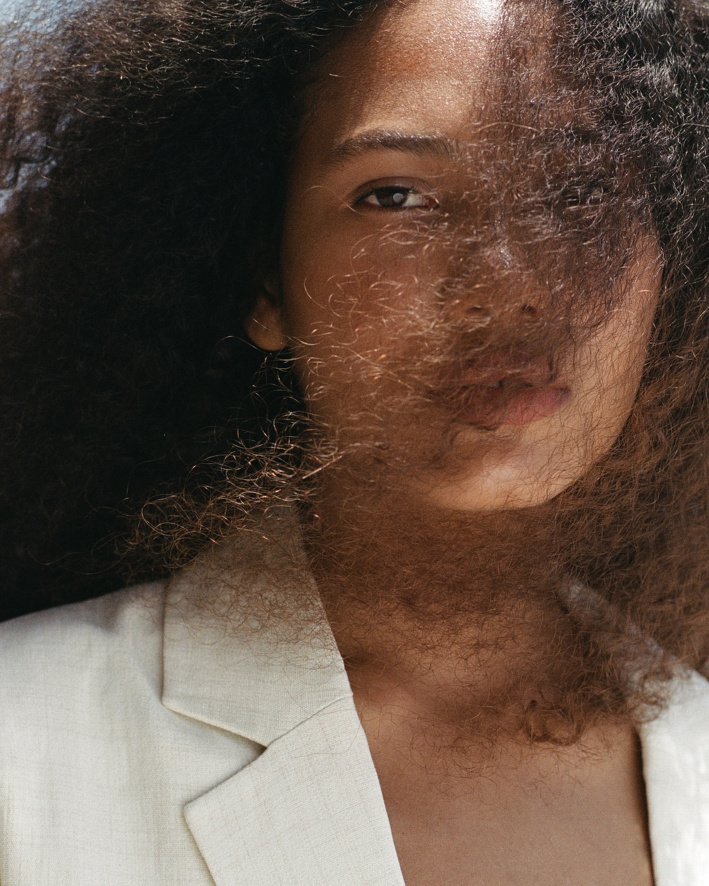 Close-up portrait of a woman with dark curly hair falling across her face wearing a cream blazer by Claire Leahy.