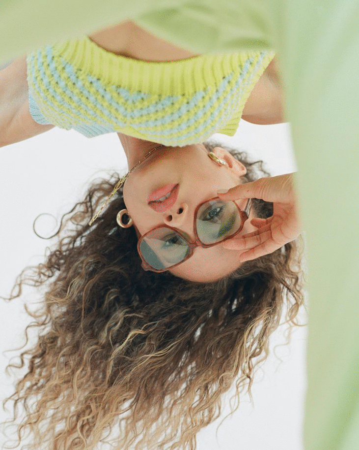 Young woman with curly hair and retro glasses poses indoors wearing a blue and yellow knit top. She is holding her glasses frame.