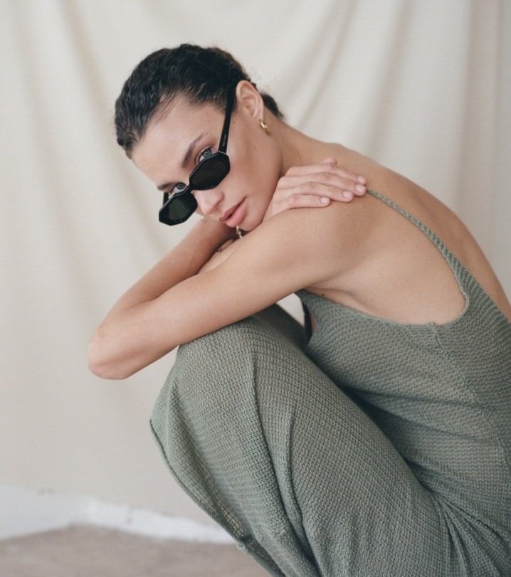 Portrait of a woman wearing sunglasses and a knitted dress, crouching against a neutral backdrop in a fashion photography style.