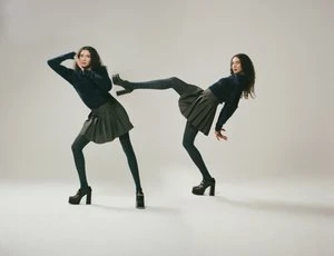 Two young women in matching outfits pose in a studio, one kicking, against a gradient white background. Fashion concept, indoor lighting.