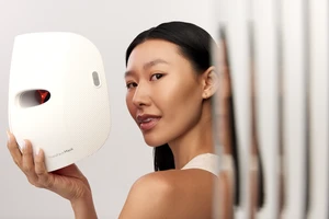 Asian woman holding a LED face mask up in a softly lit interior space, promoting skincare and facial treatments, focus on beauty.