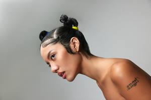 Close-up studio portrait of a young adult female looking off-camera with tied-up hair and a bare shoulder against a neutral grey background.