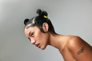 Close-up studio portrait of a young adult female looking off-camera with tied-up hair and a bare shoulder against a neutral grey background.