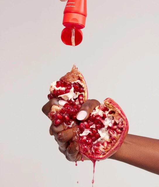 Woman squeezing a pomegranate with a tube dripping clear serum, set against a soft grey background, highlighting beauty and skincare.