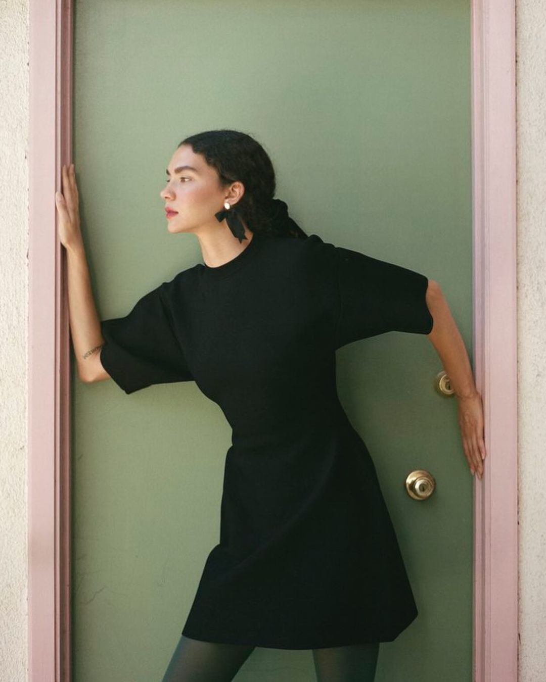 A woman in a black dress is framed by a doorway. She looks to the side, with her arm up. The door is pale green and pink.