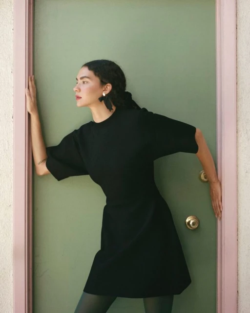A woman in a black dress is framed by a doorway. She looks to the side, with her arm up. The door is pale green and pink.