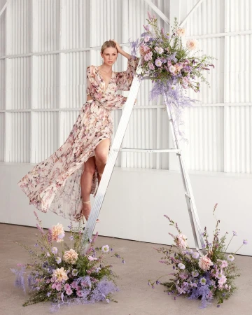 Woman in a floral print maxi dress posing on an aluminum ladder decorated with pastel floral arrangements in Los Angeles