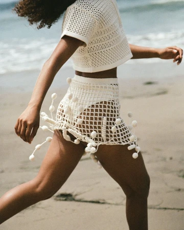 Woman running on a sandy beach wearing a cream crochet top and matching skirt with decorative fringing.