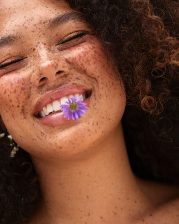 Close-up portrait of a woman with freckles and dark curly hair holding a purple flower between her teeth