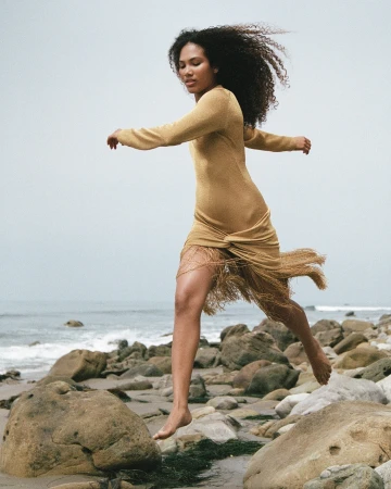 Woman with long curly hair wearing a gold fringe dress leaping across a rocky beach by the ocean, photographed by Claire Leahy
