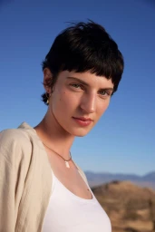 Close-up portrait of a woman with short dark hair wearing a white tank top outdoors against a clear blue sky.