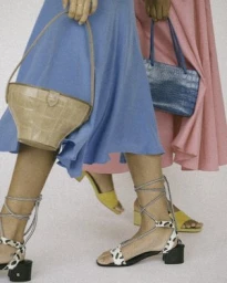 Two women holding handbags while wearing brightly coloured skirts and lace-up sandals against a plain background