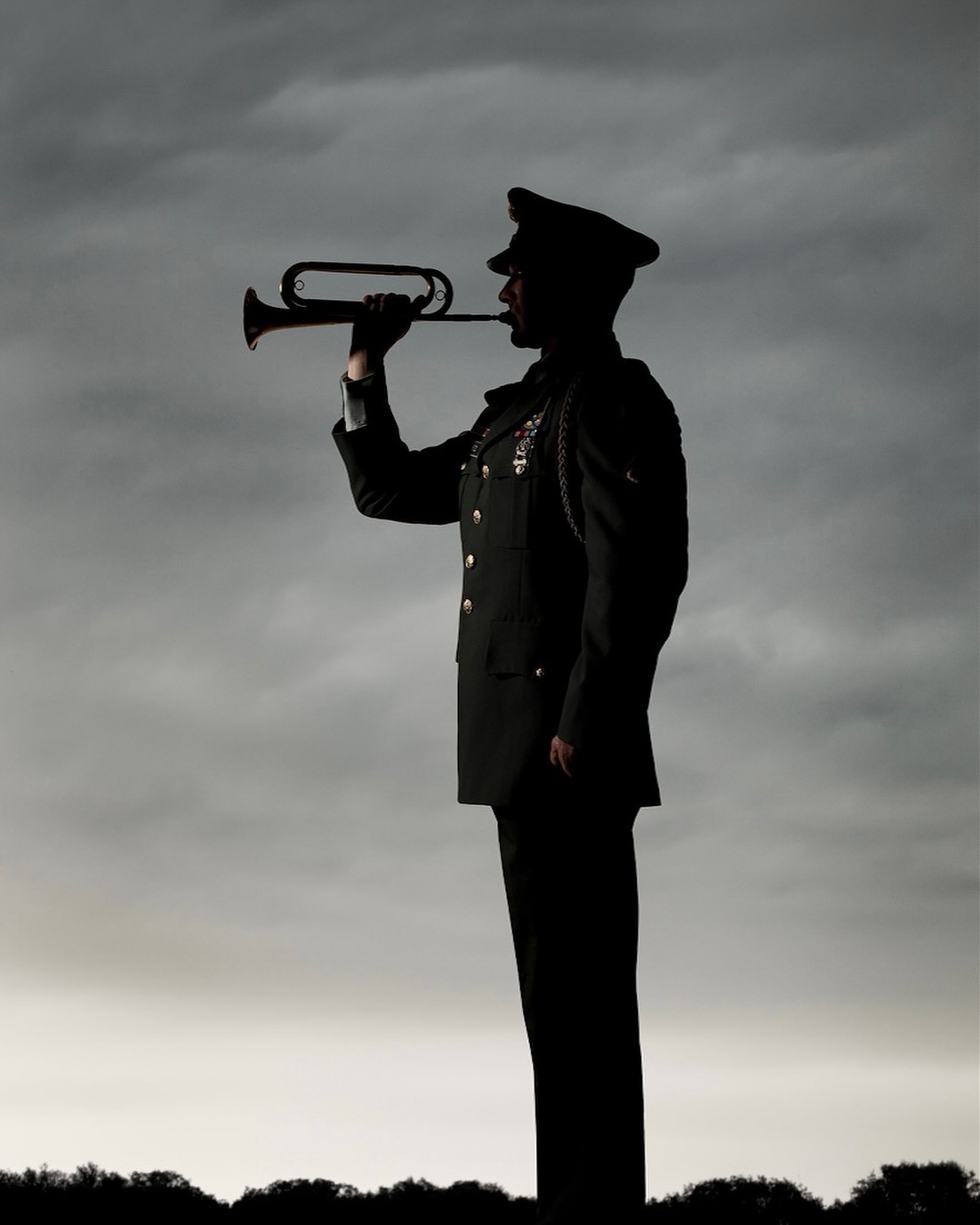 Silhouette of a uniformed person playing a bugle outdoors against a grey sky, highlighting military or formal musical performance.