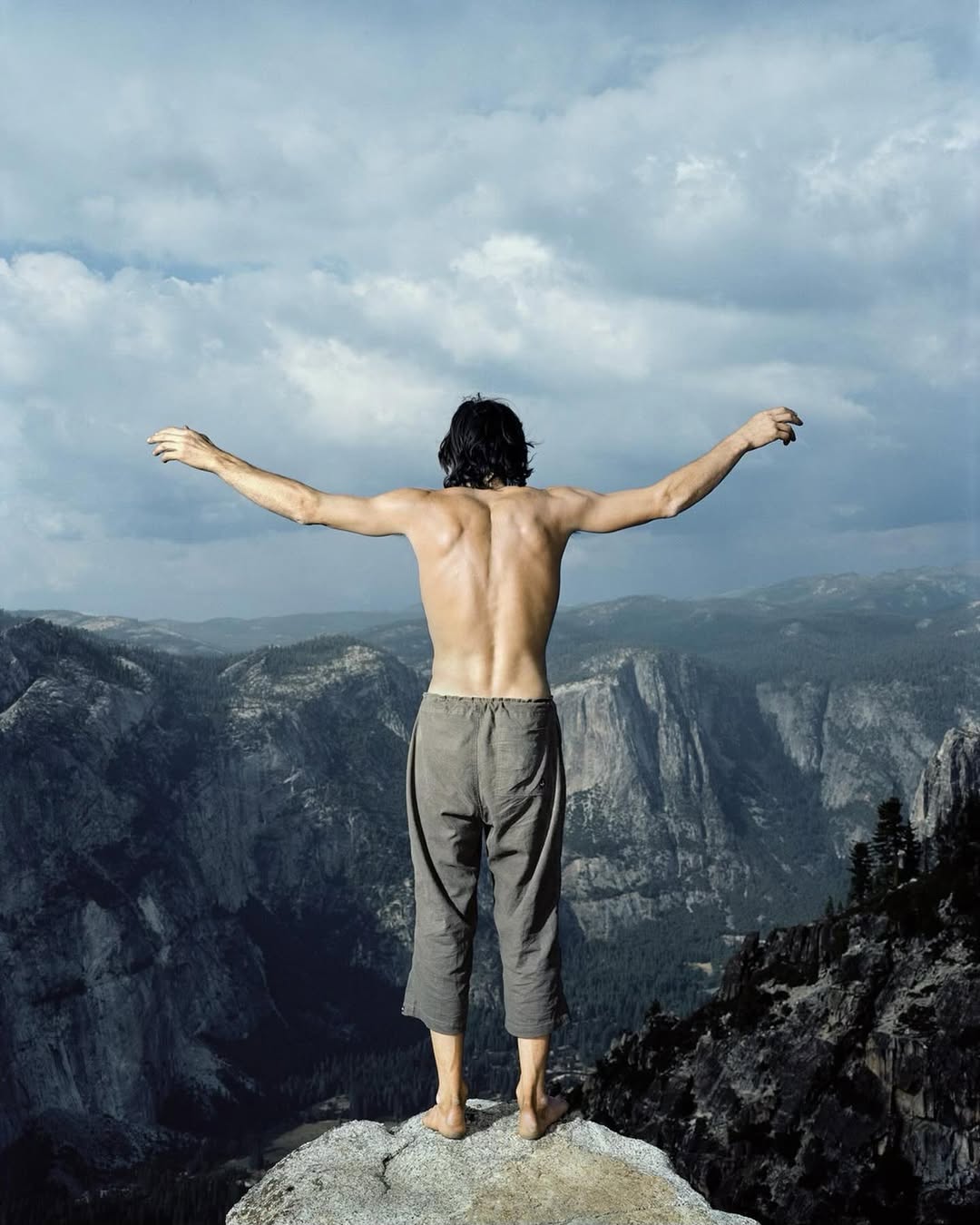A shirtless man stands on a cliff edge with arms outstretched, overlooking a vast mountain range beneath a cloudy sky in daylight.