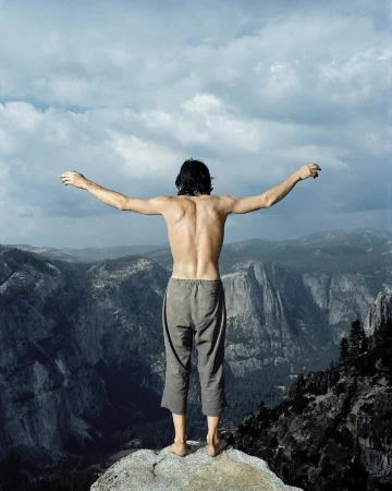 A shirtless man stands on a cliff edge with arms outstretched, overlooking a vast mountain range beneath a cloudy sky in daylight.
