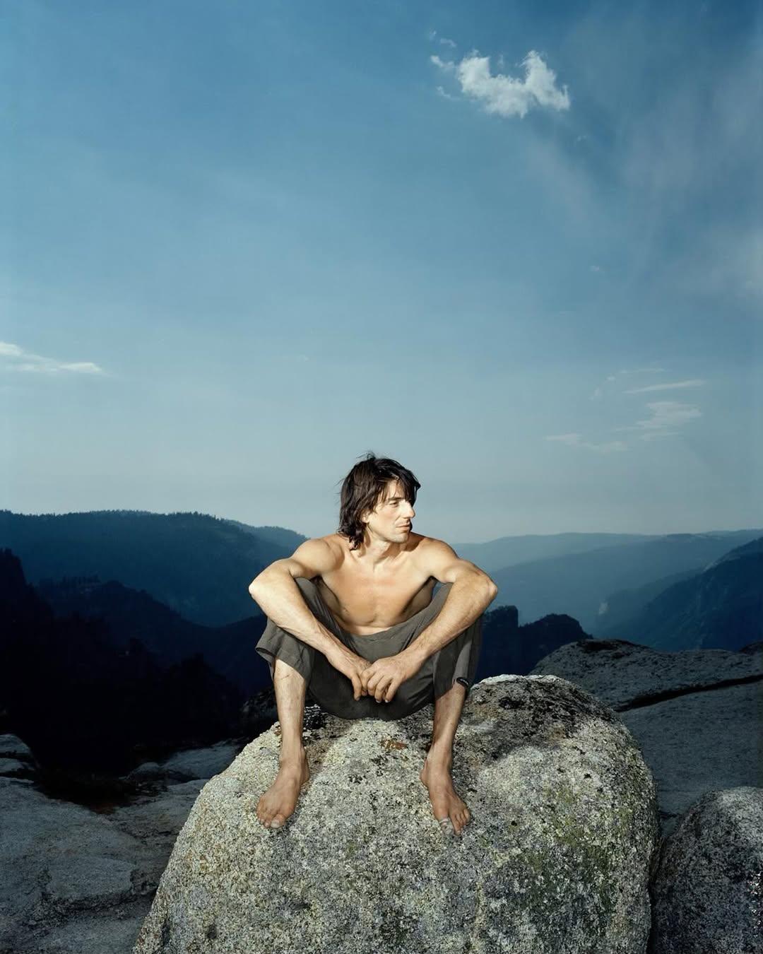 A shirtless man sits on a rock with a mountain range and blue sky in the background, creating a natural and serene outdoor scene.
