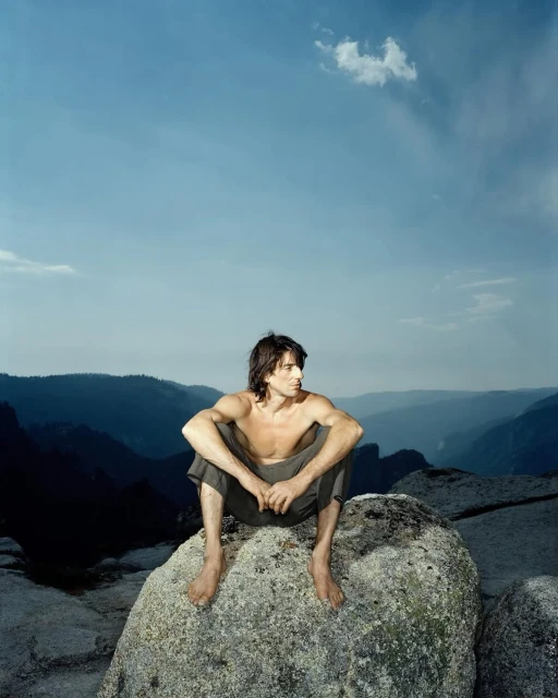 A shirtless man sits on a rock with a mountain range and blue sky in the background, creating a natural and serene outdoor scene.