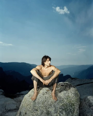 A shirtless man sits on a rock with a mountain range and blue sky in the background, creating a natural and serene outdoor scene.