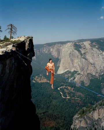 A daring shirtless man balances on a tightrope strung between two cliffs in a mountain landscape under a clear blue sky on a sunny day.