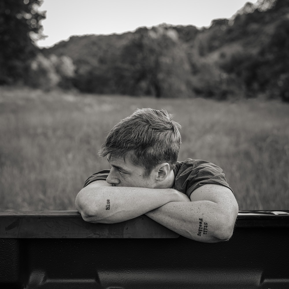 Monochrome portrait of a pensive young man resting on a truck bed, with a rural, grassy landscape and forested hills in the background.