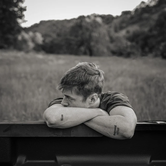 Monochrome portrait of a pensive young man resting on a truck bed, with a rural, grassy landscape and forested hills in the background.