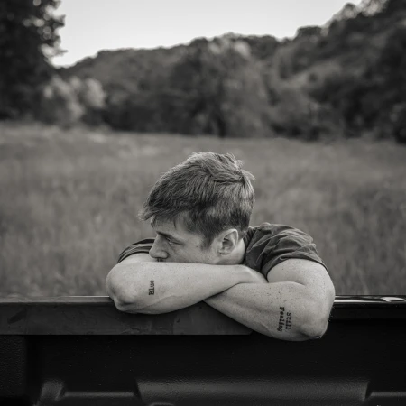 Monochrome portrait of a pensive young man resting on a truck bed, with a rural, grassy landscape and forested hills in the background.