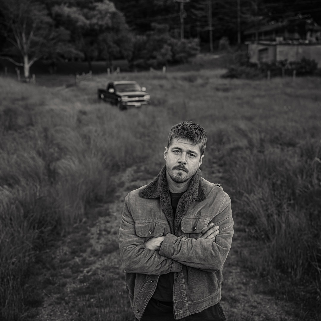 Monochrome portrait of a man with a beard, arms crossed, standing in a field with a vintage pickup truck in the background.