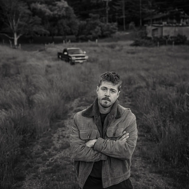 Monochrome portrait of a man with a beard, arms crossed, standing in a field with a vintage pickup truck in the background.