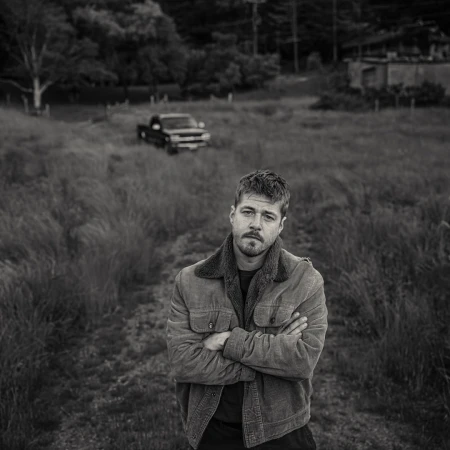 Monochrome portrait of a man with a beard, arms crossed, standing in a field with a vintage pickup truck in the background.
