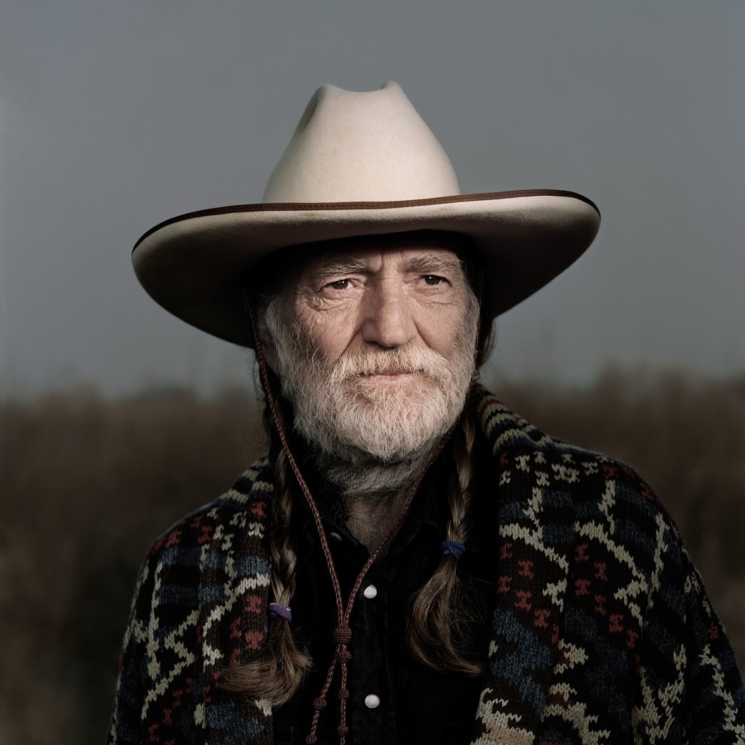 Close-up portrait of Willie Nelson wearing a cowboy hat and patterned shawl, set against a blurred outdoor background.
