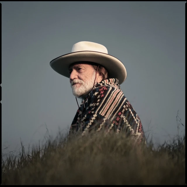 Portrait of older man wearing white cowboy hat and patterned poncho, standing in tall grass against a muted blue and gray sky.