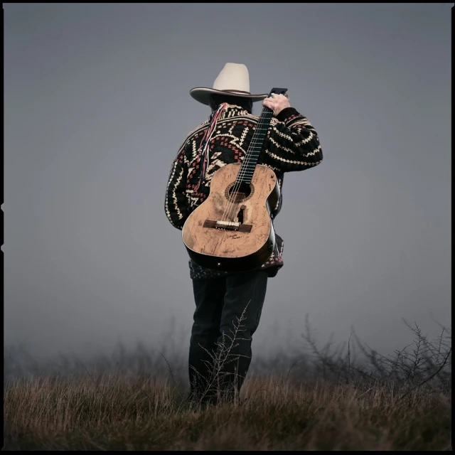 A person in a cowboy hat with their back turned is carrying a guitar in a misty, grassy landscape, evoking a country music atmosphere.