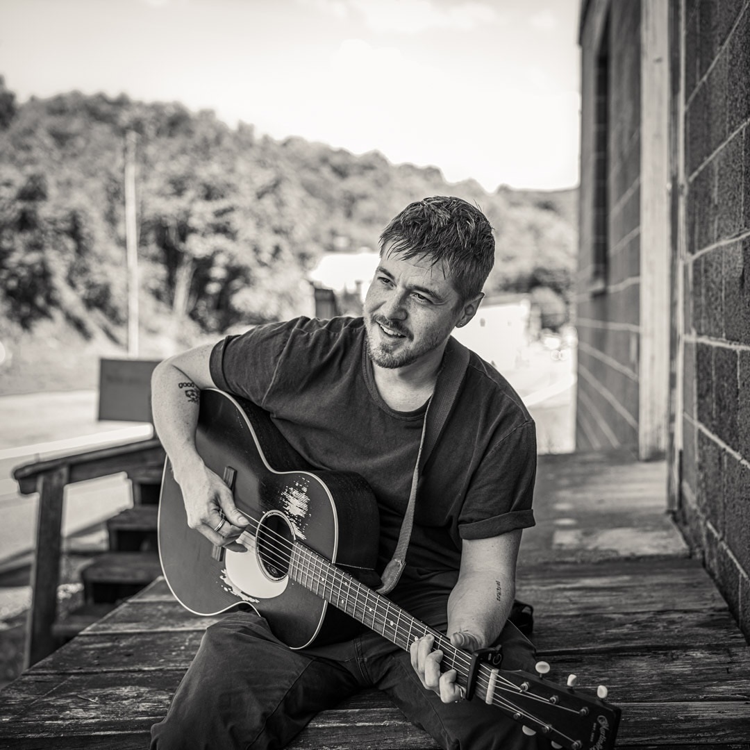 Monochrome portrait of a man with a guitar, sitting on a porch with a rural hillside in the background, creating a musical ambiance.