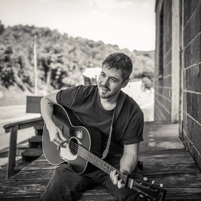 Monochrome portrait of a man with a guitar, sitting on a porch with a rural hillside in the background, creating a musical ambiance.