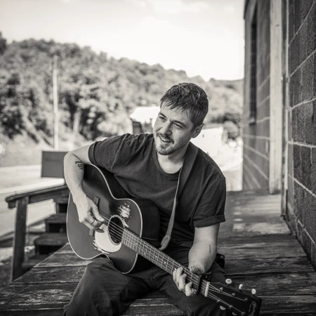Monochrome portrait of a man with a guitar, sitting on a porch with a rural hillside in the background, creating a musical ambiance.