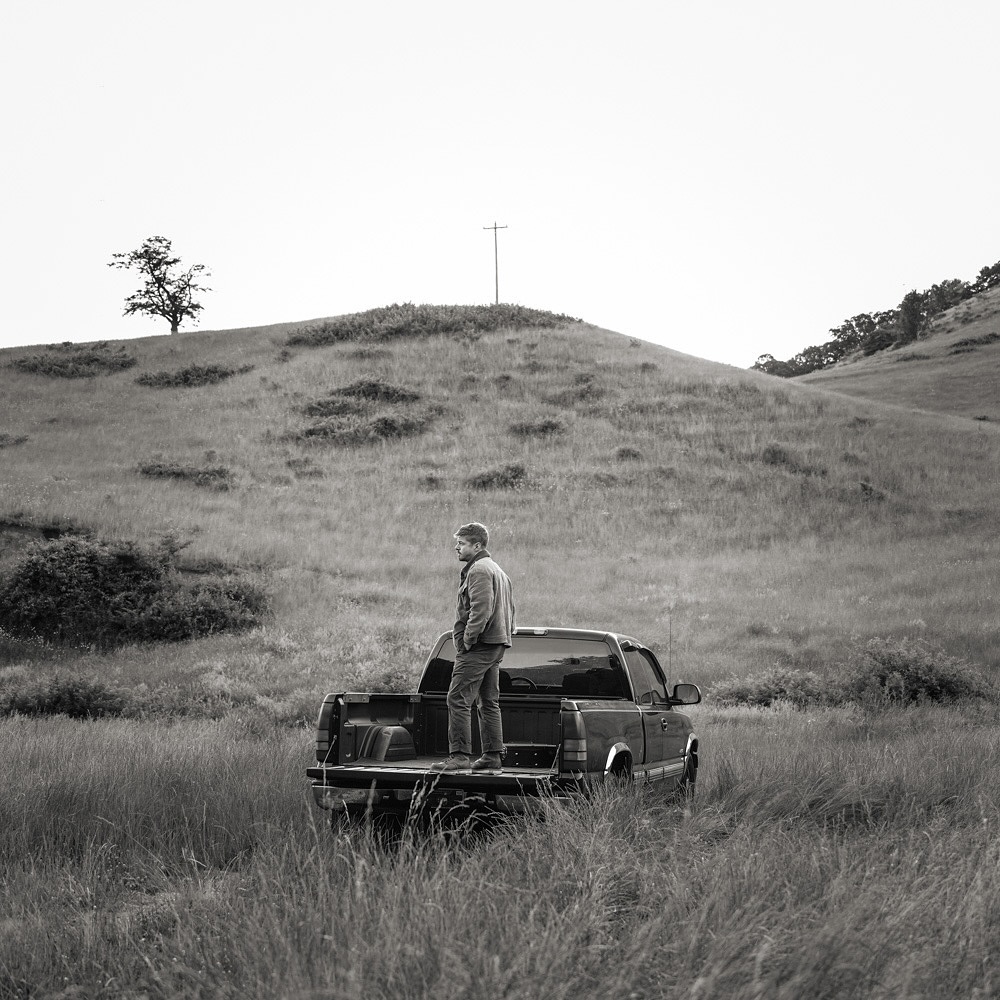 Monochrome outdoor shot of an adult male standing in the bed of a pickup truck in a grassy field, with a hill in the background.