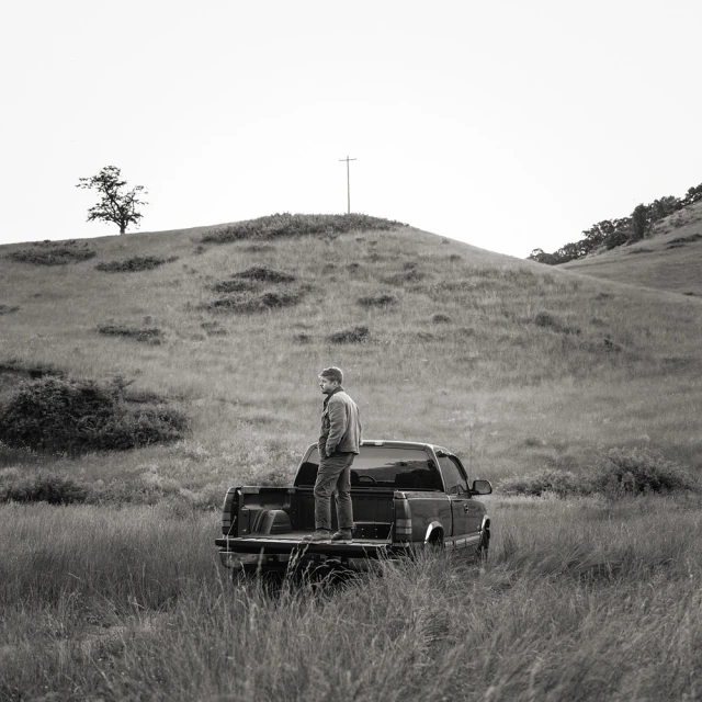 Monochrome outdoor shot of an adult male standing in the bed of a pickup truck in a grassy field, with a hill in the background.