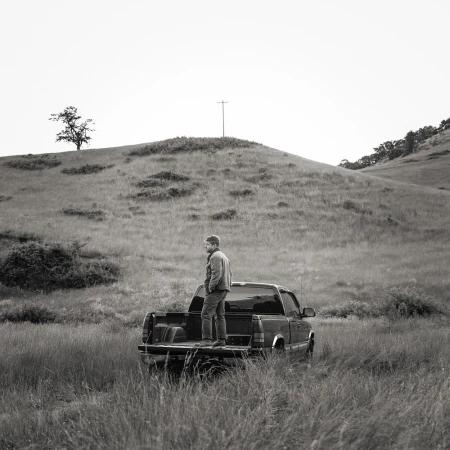 Monochrome outdoor shot of an adult male standing in the bed of a pickup truck in a grassy field, with a hill in the background.