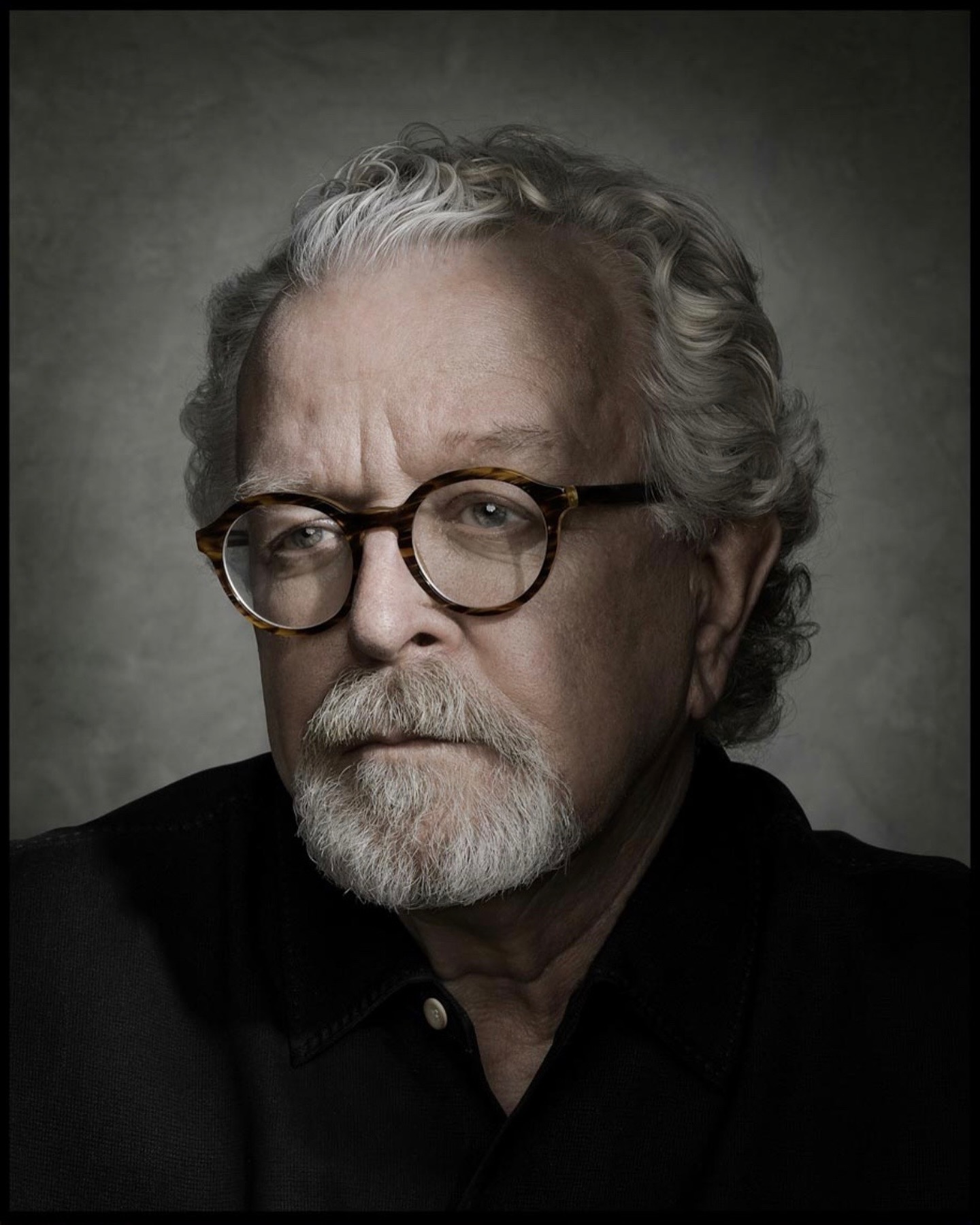 Portrait of a mature adult male with grey hair and beard wearing glasses and a black shirt against a neutral grey background.