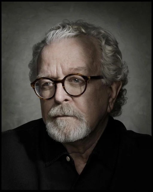 Portrait of a mature adult male with grey hair and beard wearing glasses and a black shirt against a neutral grey background.