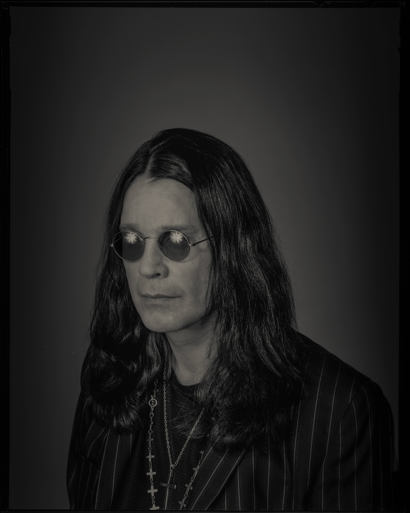 Black and white portrait of adult male celebrity, Ozzy Osbourne, wearing sunglasses, and a pinstripe blazer against a solid background.