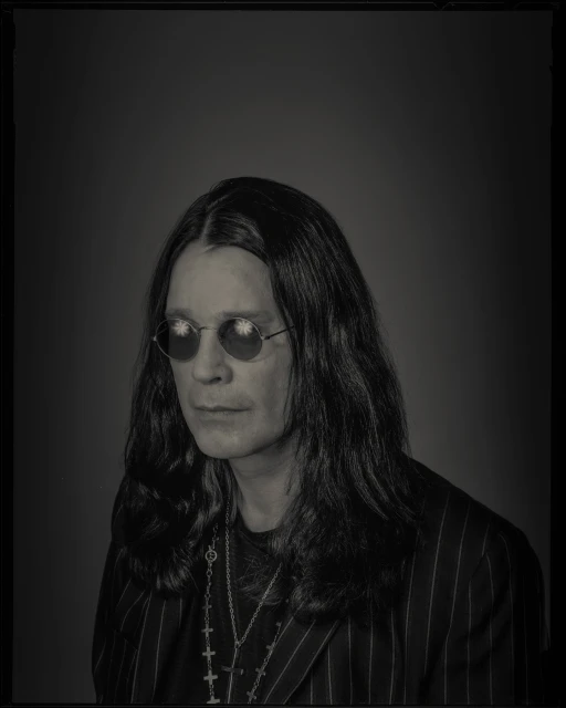 Black and white portrait of adult male celebrity, Ozzy Osbourne, wearing sunglasses, and a pinstripe blazer against a solid background.
